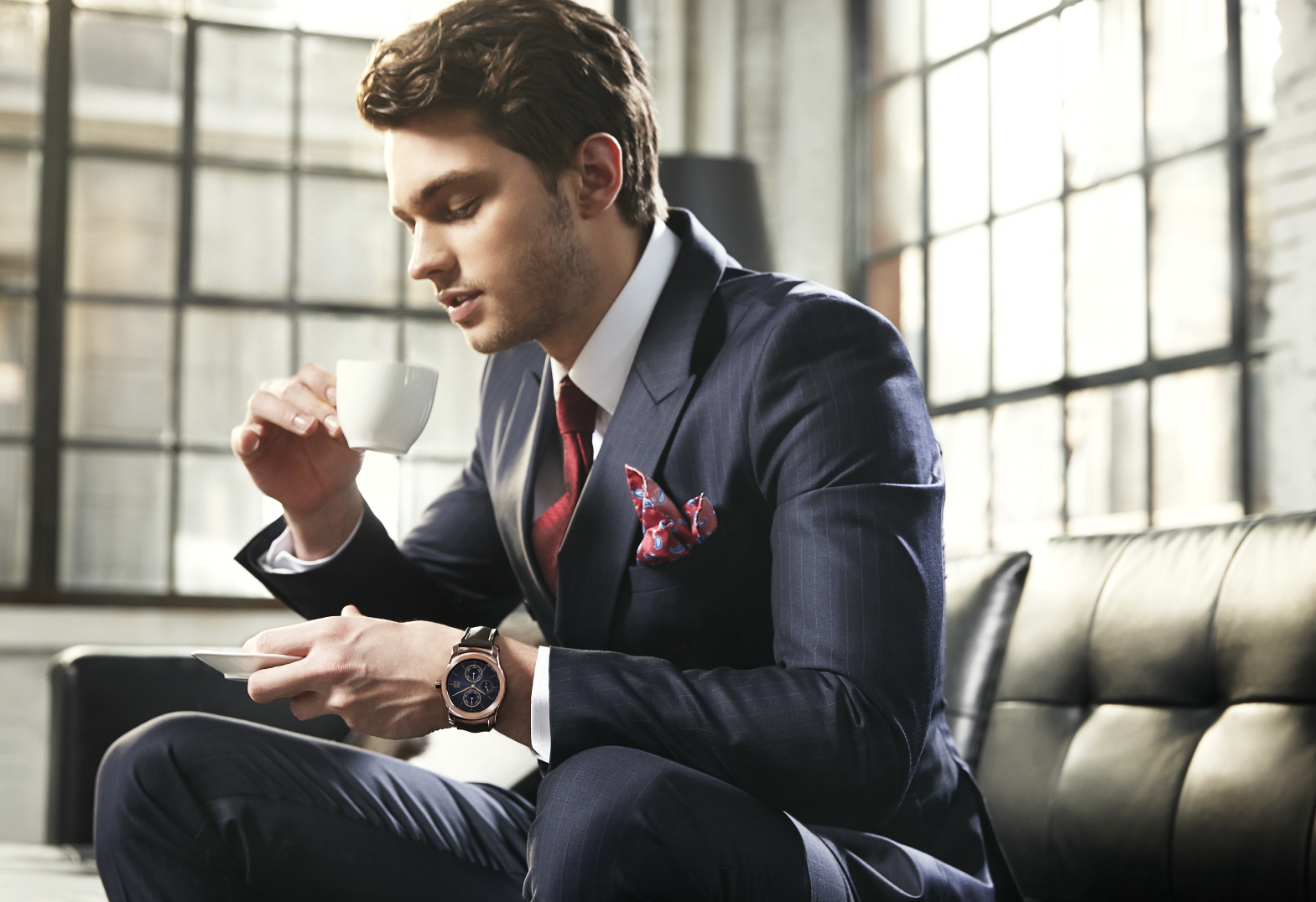 A business man in a suit wears LG's Watch Urban while drinking coffee on a couch.